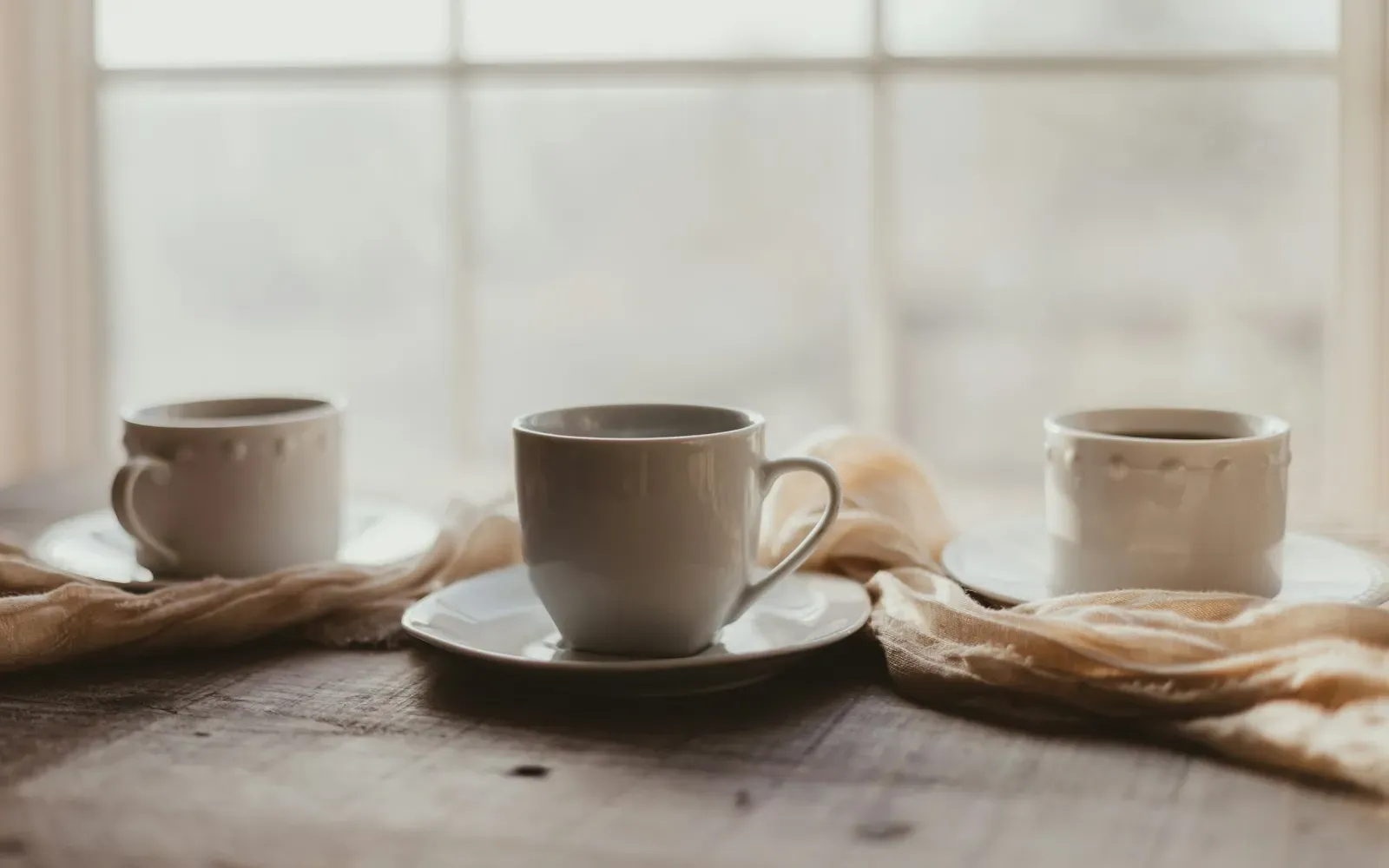 Drei Kaffeetassen auf einem Holztisch vor einem hellen Fenster